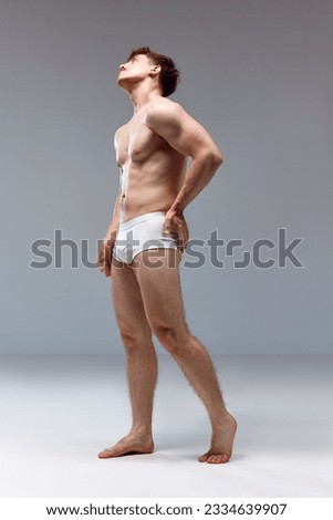 Full-length image of young man with healthy, strong, muscular body posing shirtless in underwear on grey studio background. Back pains. Concept of man's beauty, sport, health, athletic body, medicine Royalty-Free Stock Photo #2334639907