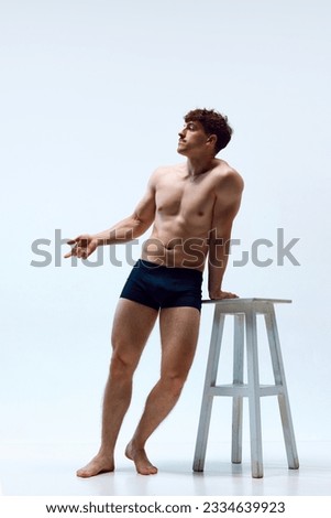 Full-length image of young man with muscular body posing shirtless, leaning on chair against white studio background. Concept of man's beauty, sportive and healthy lifestyle, athletic body Royalty-Free Stock Photo #2334639923