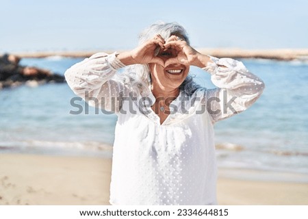 Middle age woman smiling confident doing heart gesture with hands at seaside