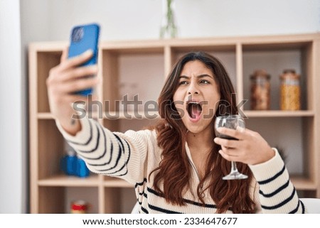 Hispanic young business woman taking a selfie picture drinking a glass of wine angry and mad screaming frustrated and furious, shouting with anger. rage and aggressive concept. 