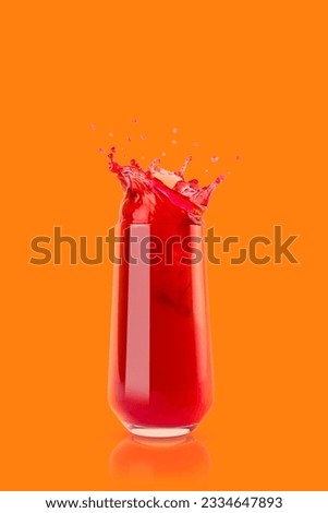 Cherry red cool juice in glass with reflection, juicy splashes and drops fly on orange background, copy space, vertical. Ripe summer fruit drink with bubbles, swirl and splashing.