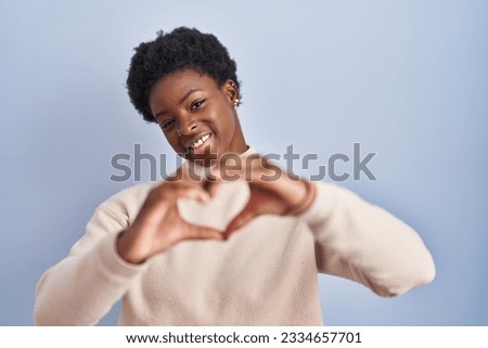African american woman standing over blue background smiling in love doing heart symbol shape with hands. romantic concept. 