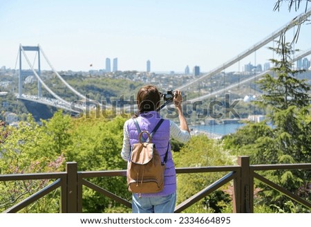A young woman tourist with a backpack photographs the cityscape with a panoramic view of the Fatih Sultan Mehmet Bridge. Back view. Otagtepe Park.