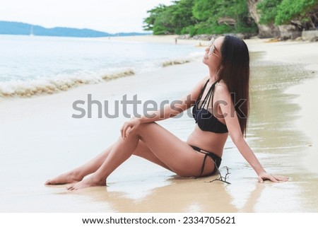 Lifestyle image of young woman wearing sunglasses wearing a black swimsuit Sitting in the sun on the beach, sea, travel, leisure time on holiday