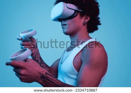 Side view of young ethnic male in VR goggles and with controllers playing video game against blue background with neon lights
