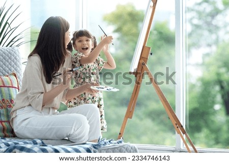 Cute little girl and mom spending good time on weekend coloring on canvas together in living room at home, beautiful green garden in the blurred background, family activity concept 