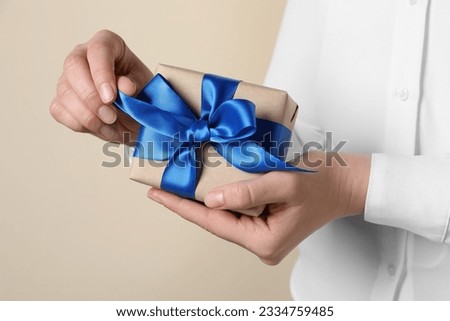Woman holding gift box with blue bow on beige background, closeup