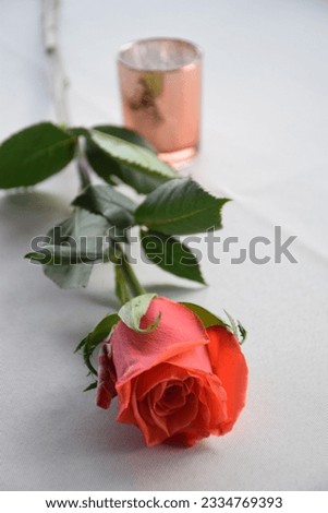 A single long stemmed red rose laying on white linen along with a pink candle holder making for a very romantic picture. 