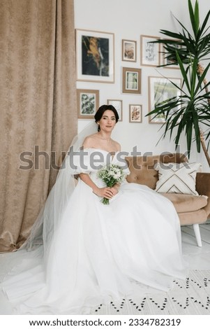 Young fashion model with perfect skin and professional makeup. A bride in a stylish dress sitting on sofa at home. Luxury wedding day. Fashion photo. Waiting for the groom. Trendy wedding style shot.