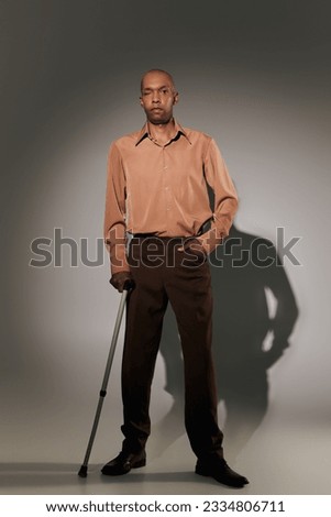 real people, bold african american man with myasthenia gravis standing with walking cane on grey background, hand in pocket, pose, dark skinned person in shirt, diversity and inclusion, full length Royalty-Free Stock Photo #2334806711