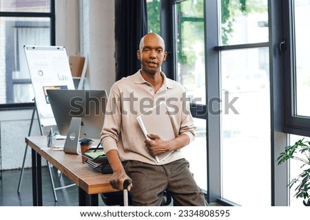dark skinned man with myasthenia gravis disease holding folder and standing with walking cane, bold african american office worker with ptosis eye syndrome, inclusion, monitor and graphics Royalty-Free Stock Photo #2334808595