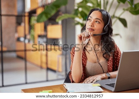 Young latin business woman wearing headphones with microphone daydreaming while working on laptop. Thoughtful mixed race businesswoman take a break and thinking about new idea.  Royalty-Free Stock Photo #2334812957
