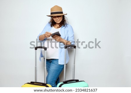 Charming multi-ethnic curly smiling pregnant woman, expectant mother with big belly in pregnancy 36 week, checking her passport and air flight ticket, standing with two suitcases over white background