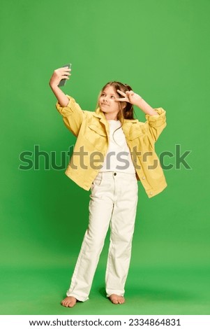 Little blogger. Full-length shot of little girl dressed stylishly taking selfie on cell phone and showing v-sign isolated on green background.