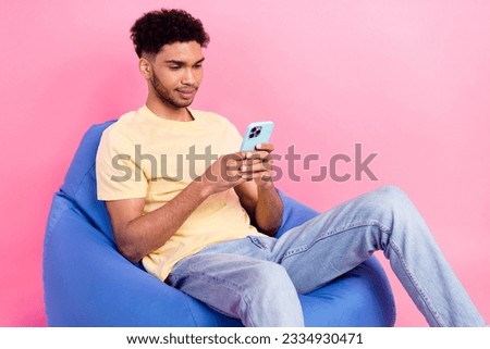 Photo of young concentrated man with wavy hair using his apple iphone last model sit beanbag rest time isolated on pink color background