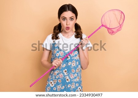 Photo of young shocked girl tails hair wear trendy overalls hunt insects hold butterfly net pouted lips isolated on beige color background