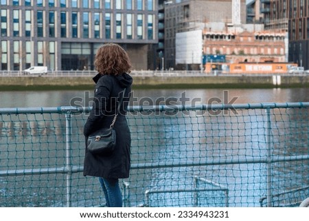 Irish girl with long red hair, dark coat and shoulder bag and blue jeans stands on the quay of the Liffey overlooking the opposite bank of Docklands in Dublin, Ireland 03-25-2023
