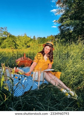 of a woman in a yellow dress and a hat painting a picture outdoors at a picnic