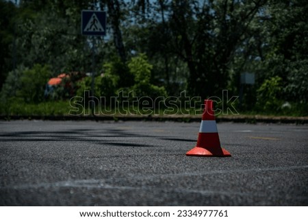 An orange traffic cone on the road. The cone stands in the middle of the road, warning of danger. The cone is made of plastic and has a trapezoidal shape. 