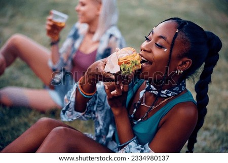 Hungry African American woman eating hamburger after open air music concert in summer.