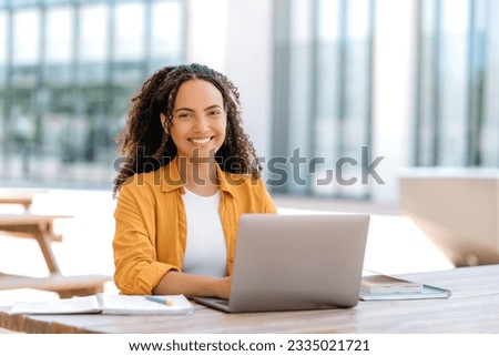 Successful woman employee. Photo of a beautiful positive brazilian or hispanic curly young woman, female student, freelancer, trader, sitting outdoors with laptop and notebook, looks at camera, smile