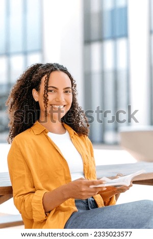 Vertical photo of a positive lovely brazilian young woman with curly hair, sitting on outdoors with a book in her hands, near the university campus, reading interesting book, looking at camera, smile