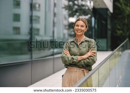Middle-aged businesswoman is standing on modern office building background and looking at camera
