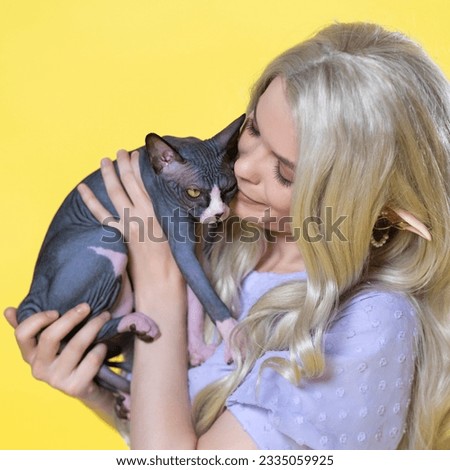Young female elf cosplay holds Sphinx kitten in her hands and gently hugs it, touching its muzzle with her lips. Elf has summer blue dress, luxurious blonde curly long hair, pearls and rings in ear.