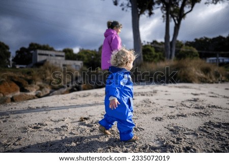 mother and baby playing on the beach. toddler in overalls walking and playing with sand and dogs on the seaside