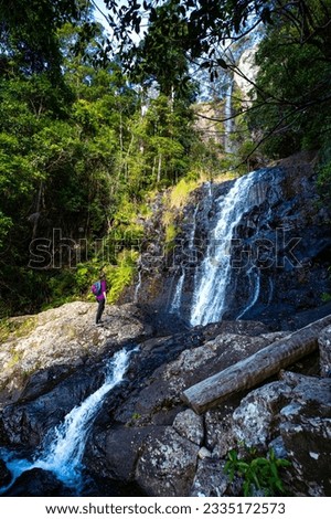 A beautiful girl stands in front of a tall, powerful waterfall; hike in Gondwana Rainforest on the Warrie Circuit trail in Springbrook National Park, Gold Coast, Queensland