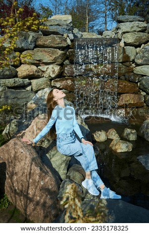A stylish beautiful young girl is sitting on a rock by a waterfall. Photo shoot in the park