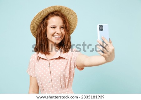 Happy young redhead curly woman 20s doing selfie shot on mobile phone post photo on social network wear casual pink dress straw hat look camera isolated on pastel blue color background studio portrait