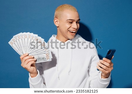 Young happy man of African American ethnicity wear white hoody hold in hand fan of cash money in dollar banknotes mobile cell phone isolated on plain dark royal navy blue background studio portrait