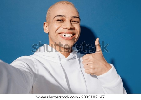 Close up young dyed blond man of African American ethnicity wear white hoody doing selfie shot pov on mobile cell phone show thumb up isolated on plain dark royal navy blue background studio portrait