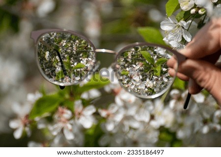 Myopia (shortsightedness) glasses in hand, looking on blooming spring flowers in focus with blurry background. Nearsighted refractive lenses outdoors in nature.