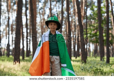 Little boy celebrating St Patrick's Day. Cute white kid dressed in a leprechaun costume and covered with large flag of Ireland