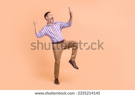 Photo of ecstatic man wear striped shirt brown trousers look at smartphone clenching fist win lottery isolated on beige color background