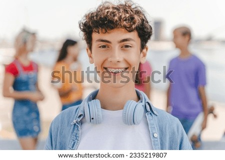 Authentic portrait of smiling teenage boy with braces wearing headphones looking at camera standing on the street with friends on background. Positive lifestyle, summer concept 