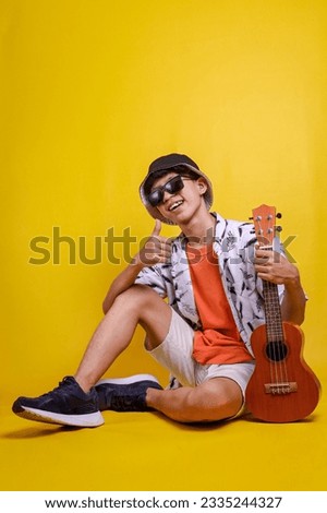 Young man wear summer clothes hold guitar ukulele and showing thumbs up while sitting on floor isolated over yellow background. Leisure, relaxation and holiday concept