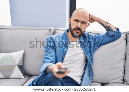 Young bald man watching tv sitting on sofa with boring expression at home