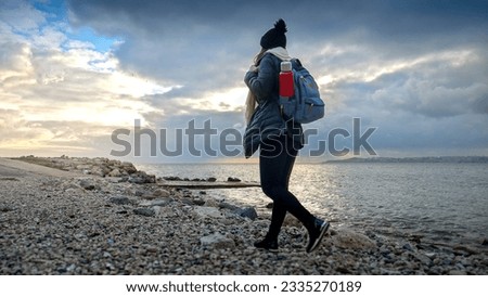 Female hiker in warm clothing equipped with a backpack and thermos, enjoying a walk on a rocky sea beach. A perfect addition to any travel or tourism project