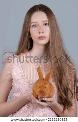 Portrait of a teenager girl with long blond hair and a red rabbit on a gray background. The girl is holding a rabbit.