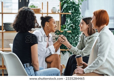 young tattooed woman holding hands of happy african american girlfriend near smiling redhead psychologist sitting with takeaway drink in paper cup, problem-solving and mental wellness concept Royalty-Free Stock Photo #2335284465