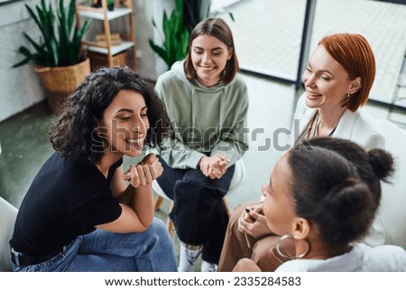 high angle view of cheerful multiracial woman talking to african american girlfriend near smiling psychologist during group therapy in consulting room, communication and mental wellness concept Royalty-Free Stock Photo #2335284583