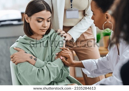 professional psychologist and african american girlfriend touching depressed and offended tattooed woman sitting with folded arms during supportive therapy, empathy and solidarity concept Royalty-Free Stock Photo #2335284613
