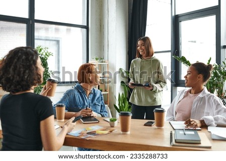 young and pleased woman standing with notebook near smiling multiethnic female friends sitting at table with coffee to go in women interest club, mutual support and understanding concept Royalty-Free Stock Photo #2335288733