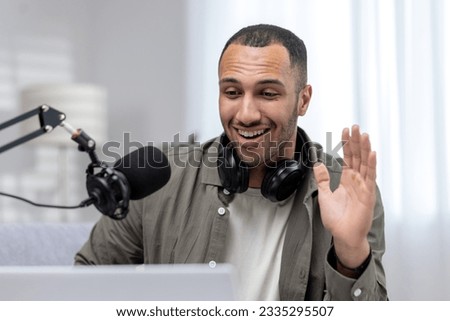 Close-up photo. Portrait of a young Latin American man working at home in a home studio. Sitting in headphones in front of a microphone, talking online through a laptop, saying hello, waving.