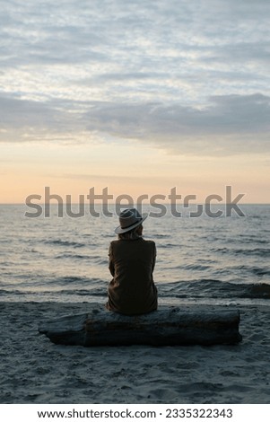 Back view of pensive lonely woman in coat and hat sitting on log on seashore looking away at sea in evening. Royalty-Free Stock Photo #2335322343