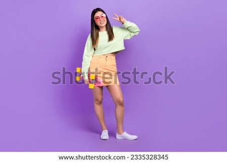 Portrait of friendly optimistic nice girl wear stylish pullover skirt hold skateboard showing v-sign isolated on violet color background
