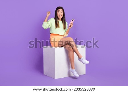 Full length photo of overjoyed crazy girl wear pullover sit on platform hold smartphone win bet isolated on violet color background